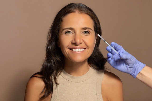 Beautician in gloves makes injection to smiling caucasian elderly woman, isolated on beige studio background. Beauty care, anti-wrinkle treatment, anti-aging restoration procedures, lifestyle